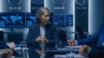 Woman wearing blazer sits at a glass table with men in suits. They are in serious discussion with many world clocks in the background and computer monitors showing maps.