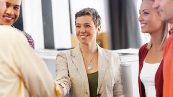 Woman wearing beige blazer and silver tear drop shaped necklace, with short greying hair, smiles as she shakes an older man's hand with other people surrounding them in an office setting.
