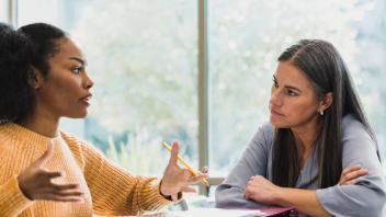 2 women sit at table together talking, woman in gold sweater gestures holding a pencil.