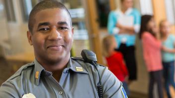 A man wearing a law enforcement uniform with a radio on his shoulder smiles at the camera while children gather in the background.