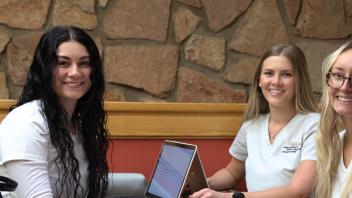 3 UCCS nursing students in white scrubs sit at table in front of laptops.