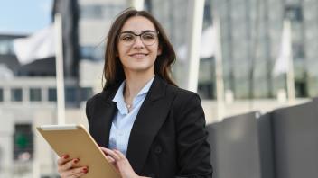 Woman smiling, wearing glasses and blazer with tablet in her hands. She stands in front of flags, with buildings in the background.