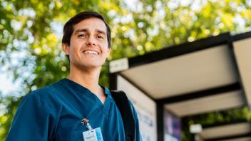 Nurse man with official id pass clipped to dark teal scrub shirt smiles in front of leafy green trees.