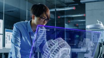 Young man with black hair and glasses, looks into a blue holographic screen with a large engineering cog system.