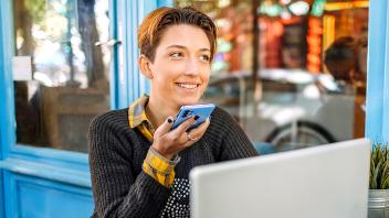 Young woman with short hair sits outside in front of a bright blue building at a table with laptop in front of her while she talks on a cell phone, smiling.