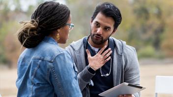 Man with dark hair wearing a grey hoodie and smart watch, holding a tablet. A stethoscope is draped around his neck. He sits outside on a bench with a woman in a jean jacket who he is explaining to by gesturing with a flat hand to his chest.
